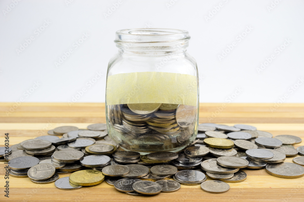 Coins in glass container with blank label on wooden surface against white background.Financial concept.Selective focus. Coins in glass container with blank label on wooden surface against white background.Financial concept.Selective focus.