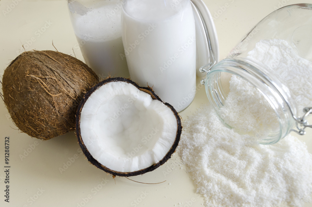 Coconut products. Cracked open coconut with meat cut in half, grounded flakes in a mason jar, flour and fresh milk in glass bottles on a table with white background and heart shaped. Coconut products. Cracked open coconut with meat cut in half, grounded flakes in a mason jar, flour and fresh milk in glass bottles on a table with white background and heart shaped.