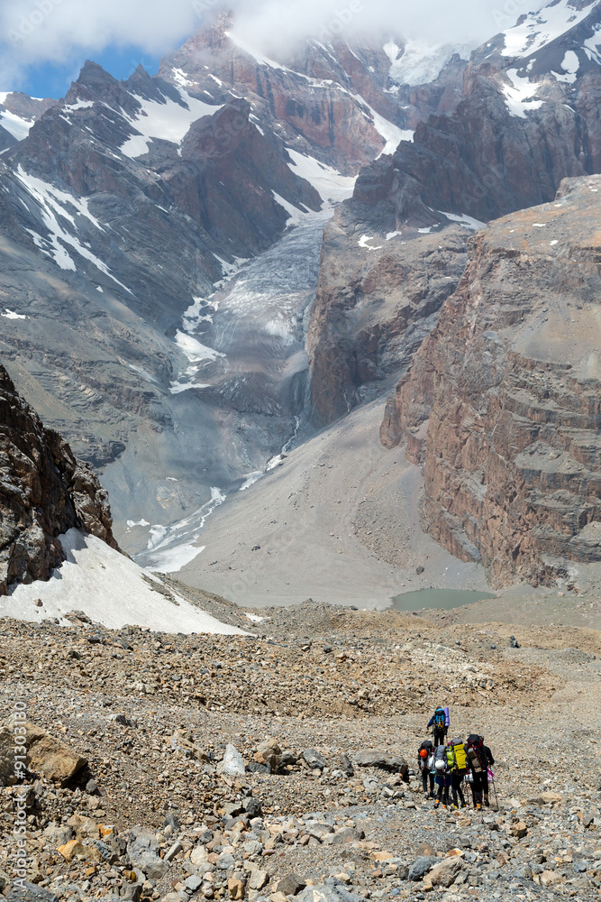 Group of people walking on rocky moraine Sport Team with Hiking Gear sporty Clothing on Mountain and Lake Landscape background High Snowbound Peaks and Massive Glacier View Behind