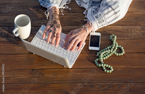Female Hands Working on Laptop Natural Wooden Desk with White Computer Large Mug Telephone and Rosary Woman Typing on Keyboard with Stylish Tattoo on Wrist Top View photo