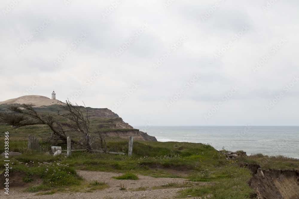 West Coast Denmark. A view to a distant lighthouse showing the erosion of the cliffs. A lone grave, dilapidated and in disrepair is in danger of falling down the cliff.