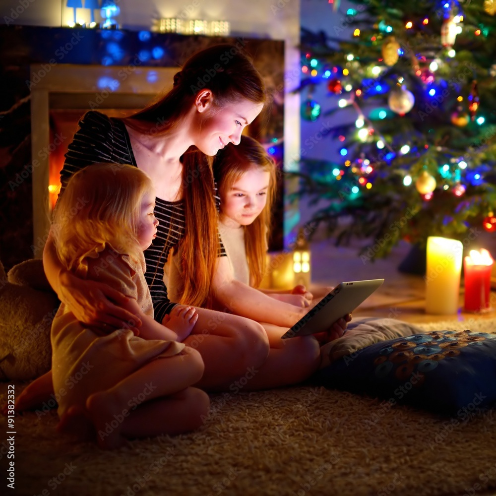 Mother and daughters using a tablet by a fireplace on Christmas Mother and daughters using a tablet by a fireplace on Christmas