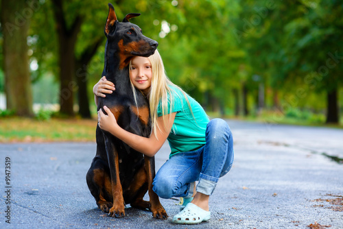 blonde girl hugs her beloved dog or doberman in summer park