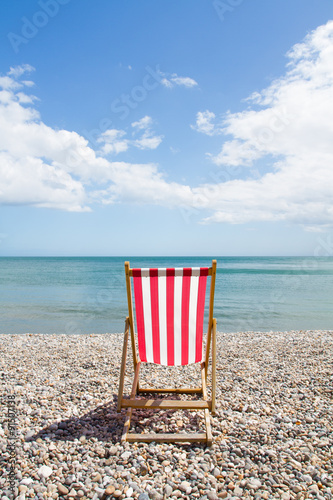 Photo A single red, striped deckchair sits on a pebble beach on a sunny day