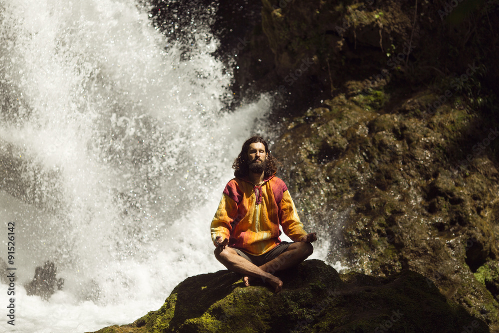 Young man meditating next to waterfall in the mountains - obrazy, fototapety, plakaty 