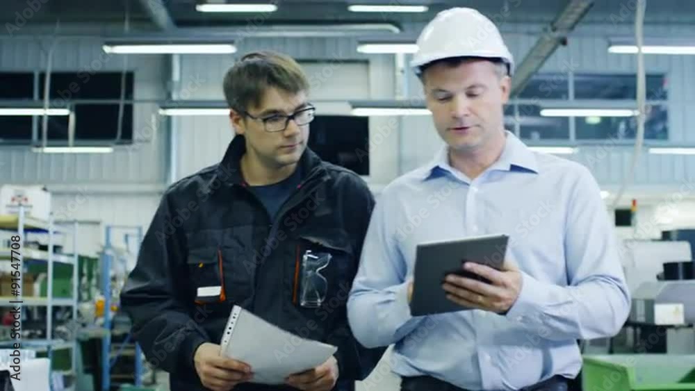 Engineer in Hard Hat and Factory Worker are Walking through Production Facility Engineer in Hard Hat and Factory Worker are Walking through Production Facility