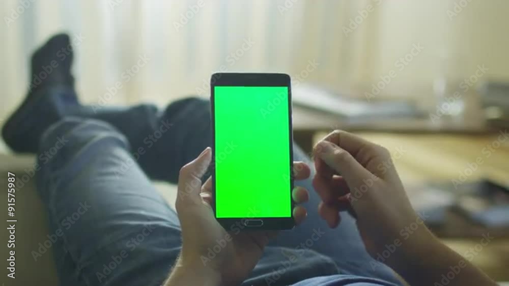 Man is Laying on Couch at Home and Using Android Phone with Green Screen in Portrait Mode on Lap. Man is Laying on Couch at Home and Using Android Phone with Green Screen in Portrait Mode on Lap.