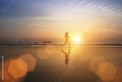 Silhouette of a young jogger girl running through the surf at amazing sunset.