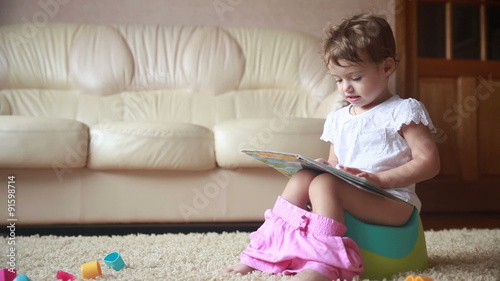 Clever toddler girl sitting on a potty and reading book.