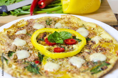 A spinach, chili, mint and pepper omelette in a dish over bamboo table cloth and chopping board with knife and raw vegetables on while background. A healthy diet meal omelet. photo