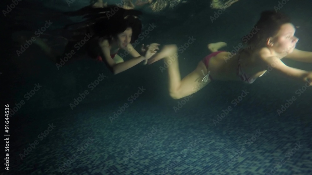 Two friends, sisters having fun dive swimming underwater in dark pool. UHD 4K stock footage