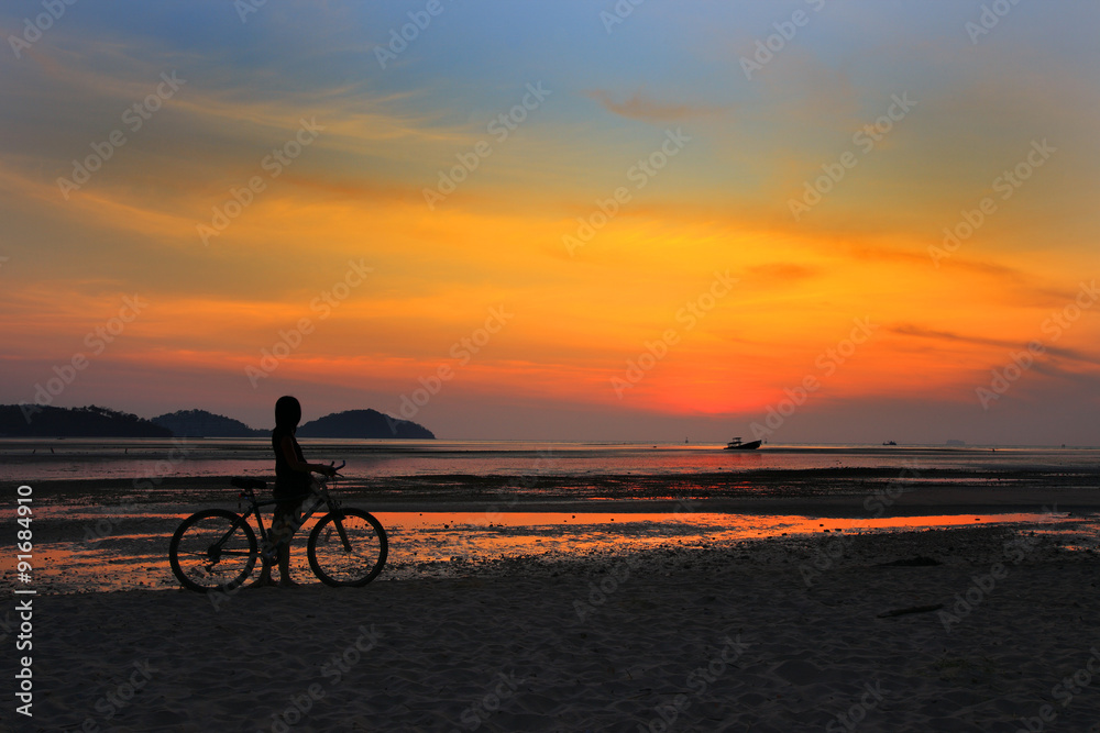 Fototapeta premium Silhouette of lonely unidentified girl with bike on beach with sunset light