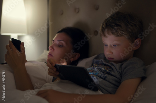  Mother with son looks in their electronic devices lying in bed