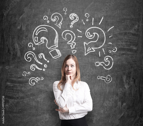 Beautiful young woman in formal clothes holds her chin and thinks about unsolved problems. Question marks are drawn around the head. Black chalk board on the background.