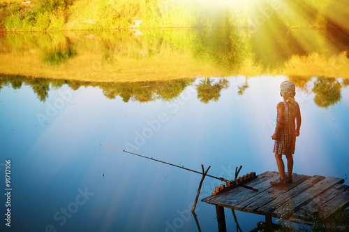 Little boy standing alone on riverside pier fishing with rod