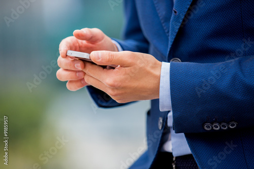 Handsome businessman in suit standing at the street with smart phone in hand. Modern skyscrapers on the background. Close-up shot
