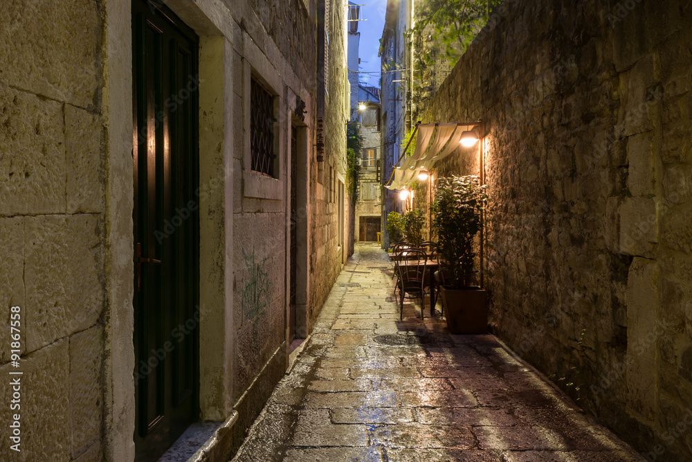 Narrow and empty alley with few cafe tables at the old town in Split, Croatia, at dark after rain.