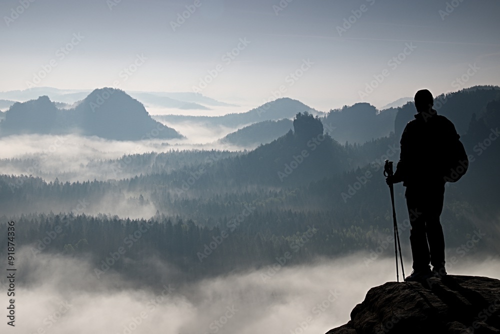 Dark silhouette of hiker with poles in hand. Sunny spring daybreak in rocky mountains. Hiker with sporty backpack stand on rocky peak  above valley.