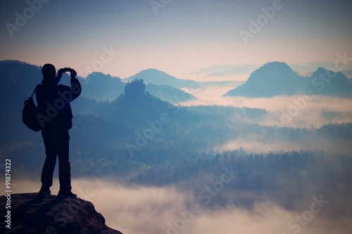 Tourist with backpack takes photos with smart phone on peak of rock. Dreamy fogy valley below