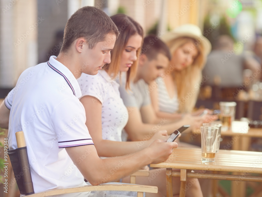 Group of teenagers sitting in a row at the sidewalk cafe using mobile phones