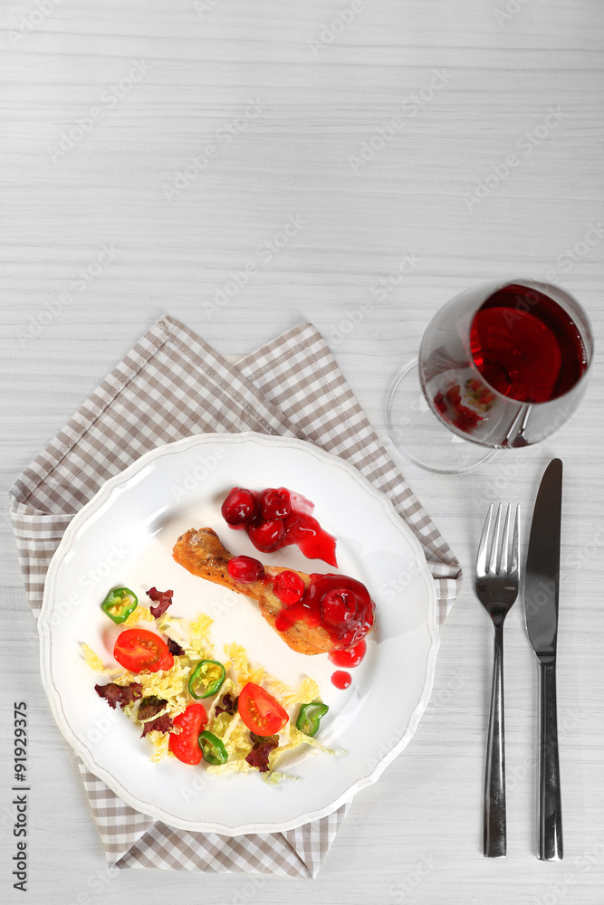 Dish of baked chicken leg and vegetable salad in white plate with glass of wine on table with napkin, top view