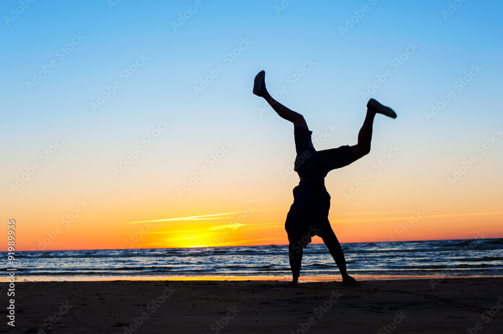 Young man making handstand on the beach. Multicolored sunset bac