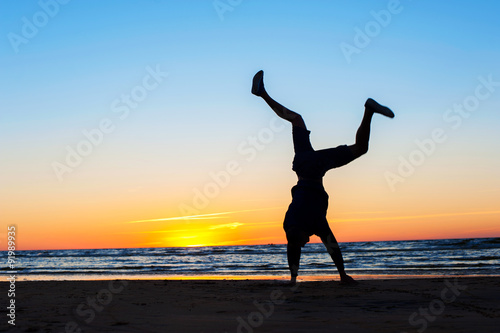 Young man making handstand on the beach. Multicolored sunset bac
