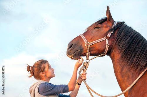 Young cheerful teenage girl stroking brown horse's nose. Outdoor