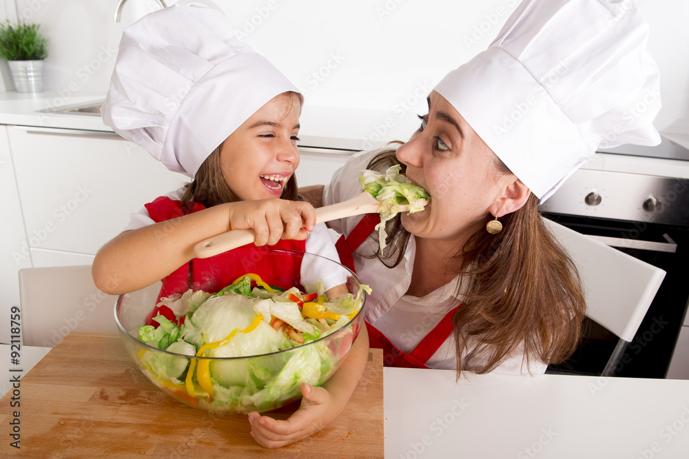 happy mother and little daughter at home kitchen preparing salad in apron and cook hat happy mother and little daughter at home kitchen preparing salad in apron and cook hat