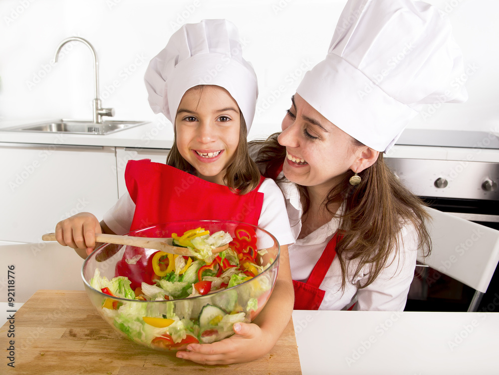 happy mother and little daughter at home kitchen preparing salad in apron and cook hat happy mother and little daughter at home kitchen preparing salad in apron and cook hat