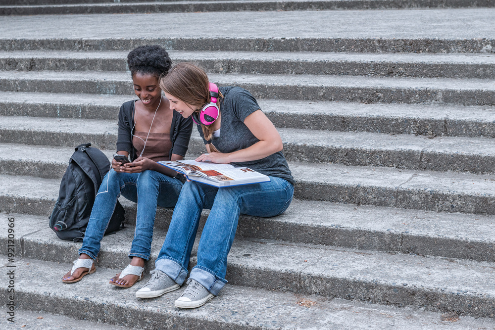 Mixed race young adult female students sitting on stairs at university interacting with smartphone