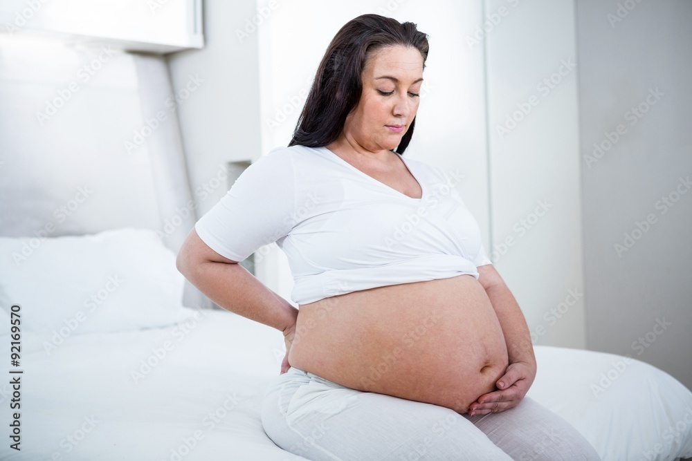 Pregnant woman holding her back while sitting on a bed Pregnant woman holding her back while sitting on a bed