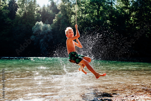 7 years boy having fun on rope swing above river.