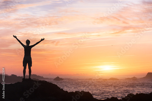 Silhouette of young woman with open arms facing the sea at sunse