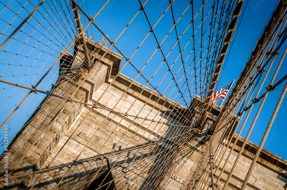 Obraz premium Closeup of the Brooklyn Bridge in New York City, USA. The American flag is flying over the cloudless blue sky
