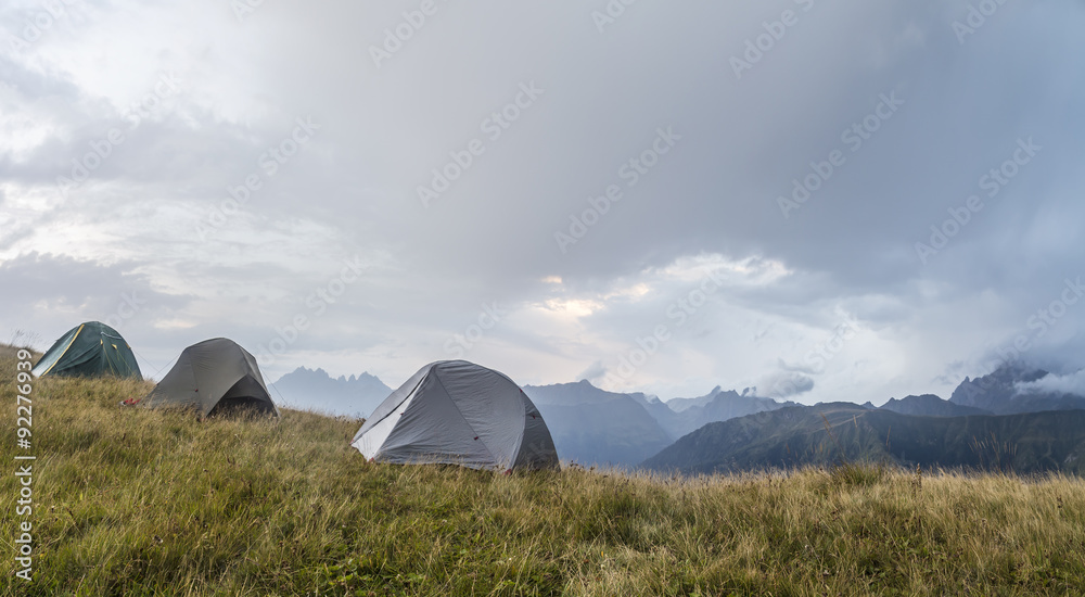 panorama of the mountains, in a clearing, there are three tents on the background of deteriorating weather and rain clouds at sunset