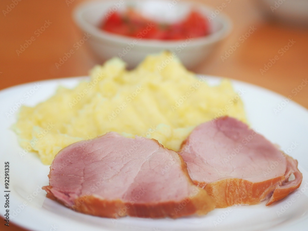 Smoked pork meat (leg) with mashed potatoes served on a white plate and a tomato salad in a bowl. Smoked pork meat (leg) with mashed potatoes served on a white plate and a tomato salad in a bowl.