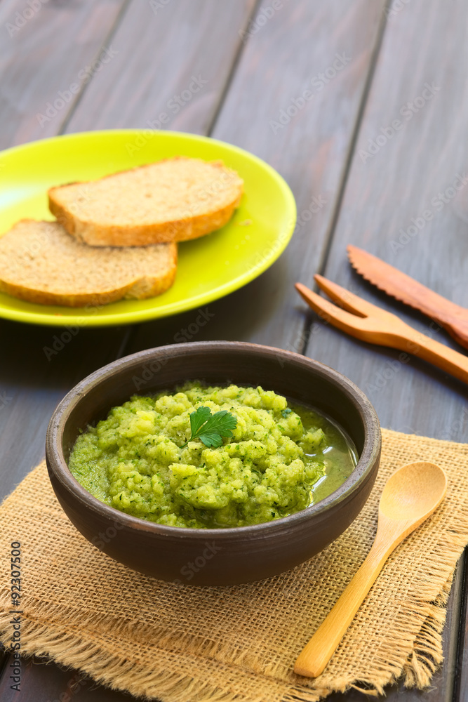 Rustic bowl of homemade zucchini and parsley spread garnished with fresh parsley leaf, slices of wholegrain bread in the back, photographed with natural light (Selective Focus, Focus on the leaf) Rustic bowl of homemade zucchini and parsley spread garnished with fresh parsley leaf, slices of wholegrain bread in the back, photographed with natural light (Selective Focus, Focus on the leaf)