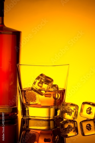 glass of whiskey with ice cubes near bottle on table with reflection, warm tint atmosphere