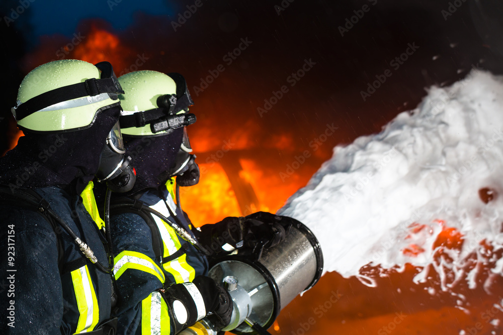 Fototapeta premium Feuerwehr - Feuerwehrmänner löschen ein großes Feuer, sie stehen vor einer Feuerwand und tragen Schutzkleidung