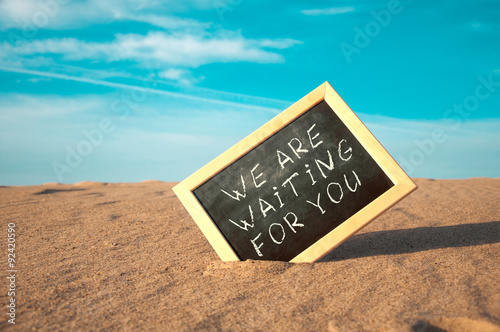 closeup of a chalkboard with a wooden frame and the word we are waiting for you written in it, placed on the sand of a beach