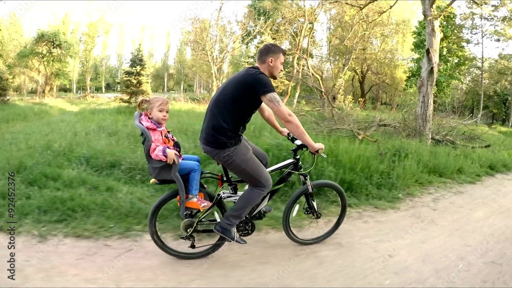 Man With Child On Backseat Riding A Bicycle In Park Man With Child On Backseat Riding A Bicycle In Park