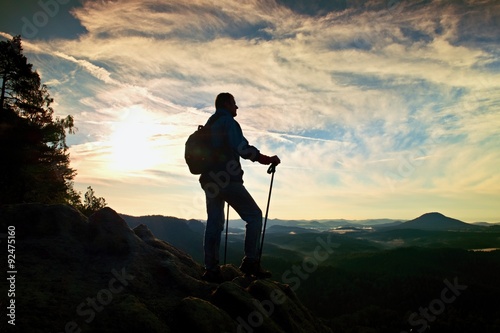 Silhouette of alone hiker with poles in hand. Tourist with sporty backpack stand on rocky view point above misty valley. Sunny spring daybreak in rocky mountains.