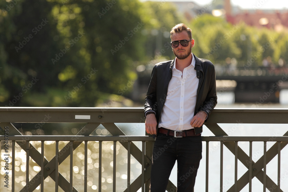 Attractive and laid back man leaning on a bridge with water behind on a sunny summer day. Wearing a leather jacket and white shirt.