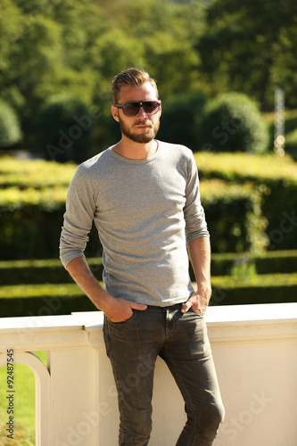 A modern man wearing a grey shirt and jeans with sunglasses, leaning back outside on a sunny summer day.