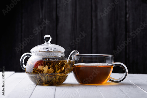 Glass teapot with blooming green tea and cup of tea on white wooden table and dark background still life