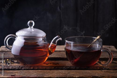 Hot tea in glass teapot and cup with spoon and steam on wooden table dark still life