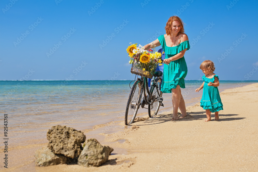 Obraz premium Happy family - baby girl with young mother after bicycle exercises walk with bike and have a fun on sea sand tropical beach. Active parents and people outdoor activity on summer vacation with children