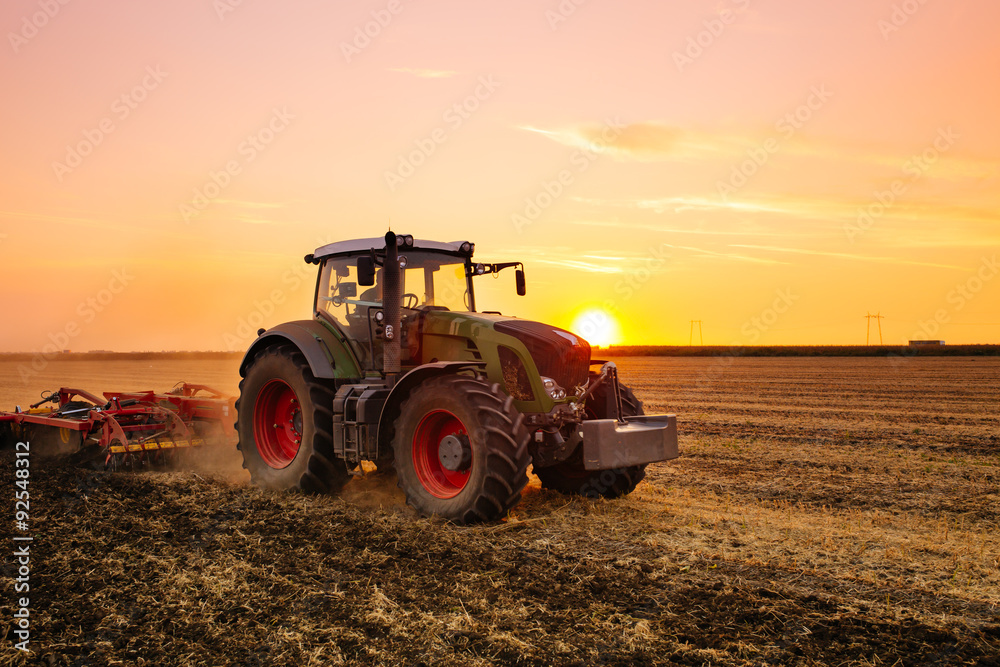 Naklejka premium Tractor on the barley field by sunset.