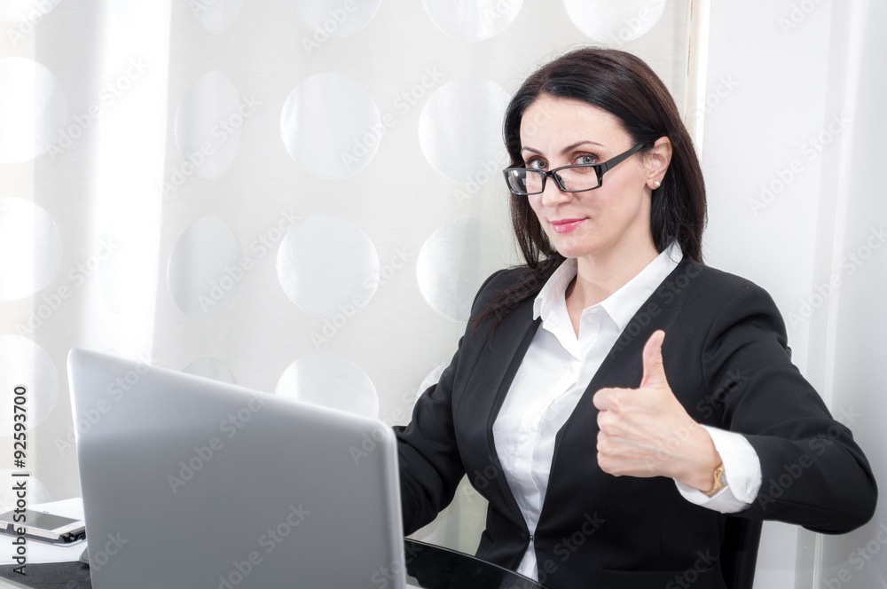 Woman working in a office shows the thumbs up gesture expressing her satisfaction with a project shown on the computer Woman working in a office shows the thumbs up gesture expressing her satisfaction with a project shown on the computer